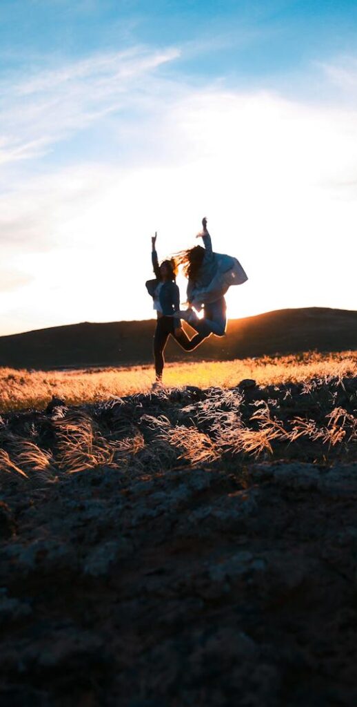 Two friends jump joyfully at sunset in a rocky field, creating a silhouette against the vibrant sky.